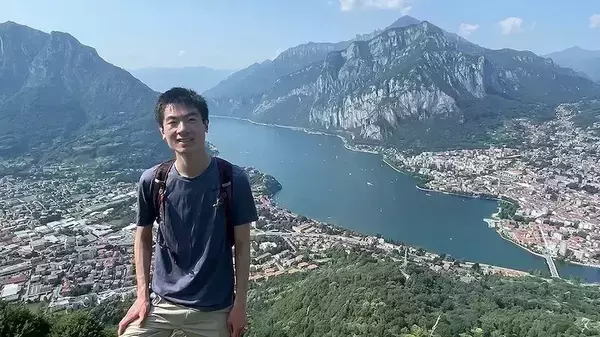 A hiker, Bryan Fok, smiles for the camera from a mountaintop overlooking the city of Lecco, Italy, and the southeastern branch of Lake Como. The sparkling blue water of the lake is dotted with boats, and the city stretches along its shores, nestled among lush green hills and towering, rocky mountains.