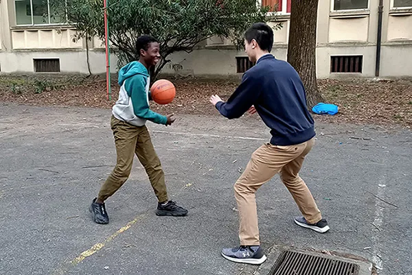 Two students play basketball on an outdoor court. One holds the ball, smiling at the other student who is guarding him.  They are both wearing khaki pants and athletic shoes.