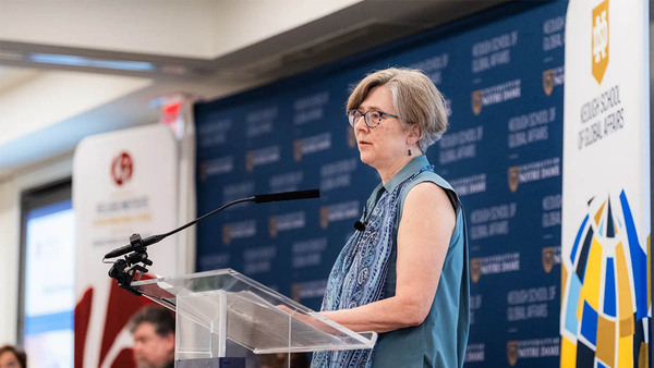 A woman with short gray hair, wearing a blue sleeveless top with a paisley print, speaks at a clear podium in front of a Keough School of Global Affairs banner.