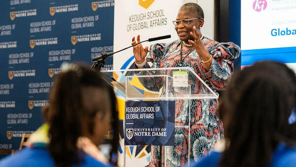 A woman gestures with her hands while speaking at a clear podium bearing the Keough School of Global Affairs logo at the University of Notre Dame.  She wears glasses, a pearl necklace, and a colorful dress with swirling red and blue patterns.  Audience members are slightly blurred in the foreground.  A branded backdrop displays the Keough School and Notre Dame logos.