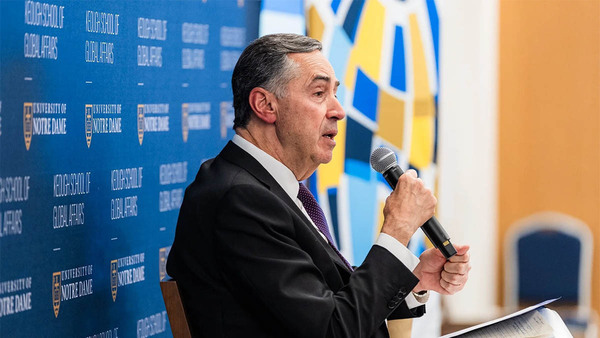 A man in a dark suit and purple tie speaks into a microphone in front of a Keough School of Global Affairs backdrop at the University of Notre Dame.