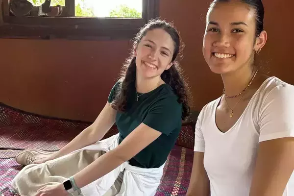 Two smiling students sit on patterned mats inside a room with a window. One student wears a dark green shirt and khaki pants, the other a white shirt.