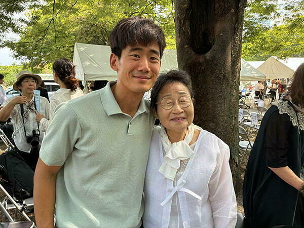 A young man in a light green polo shirt smiles as he stands next to an older woman wearing a white top with a ruffled collar.  They are posing for a photo outdoors near a tree trunk, with other people and white tents in the background.