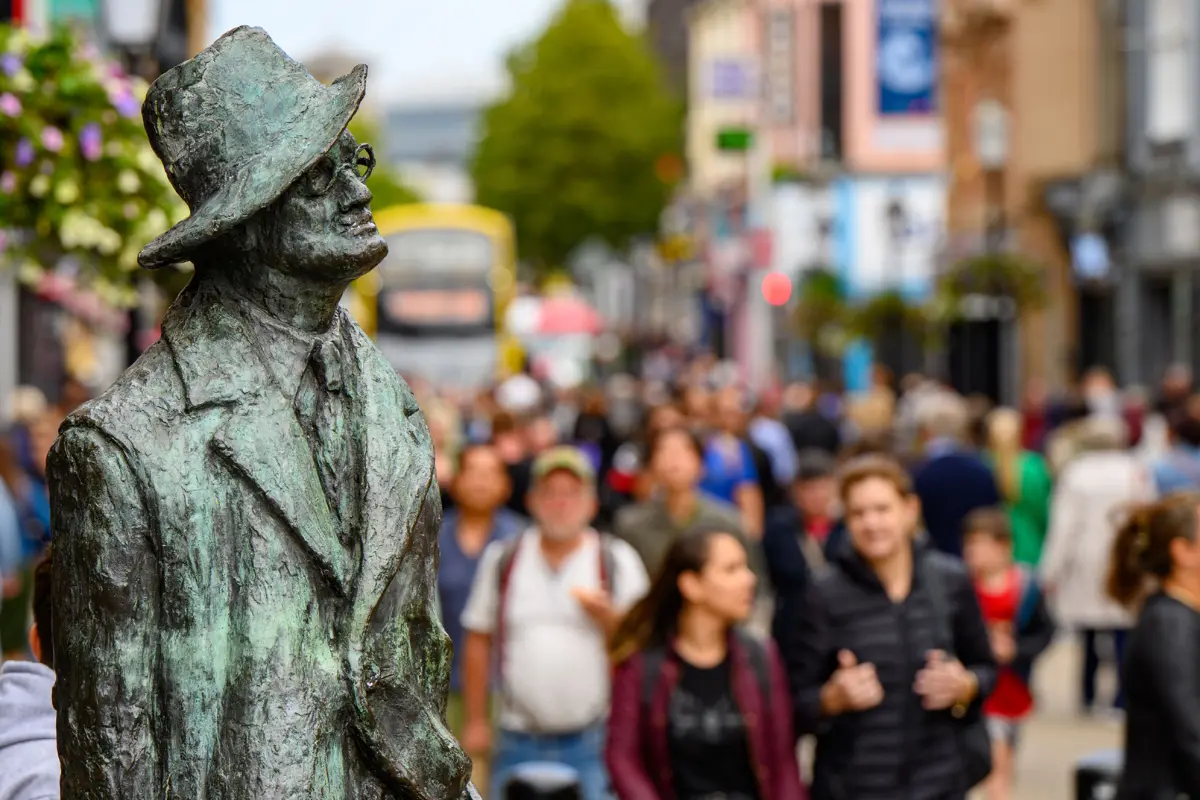 Bronze bust of James Joyce wearing a hat and glasses, gazing towards a blurred street scene filled with people.