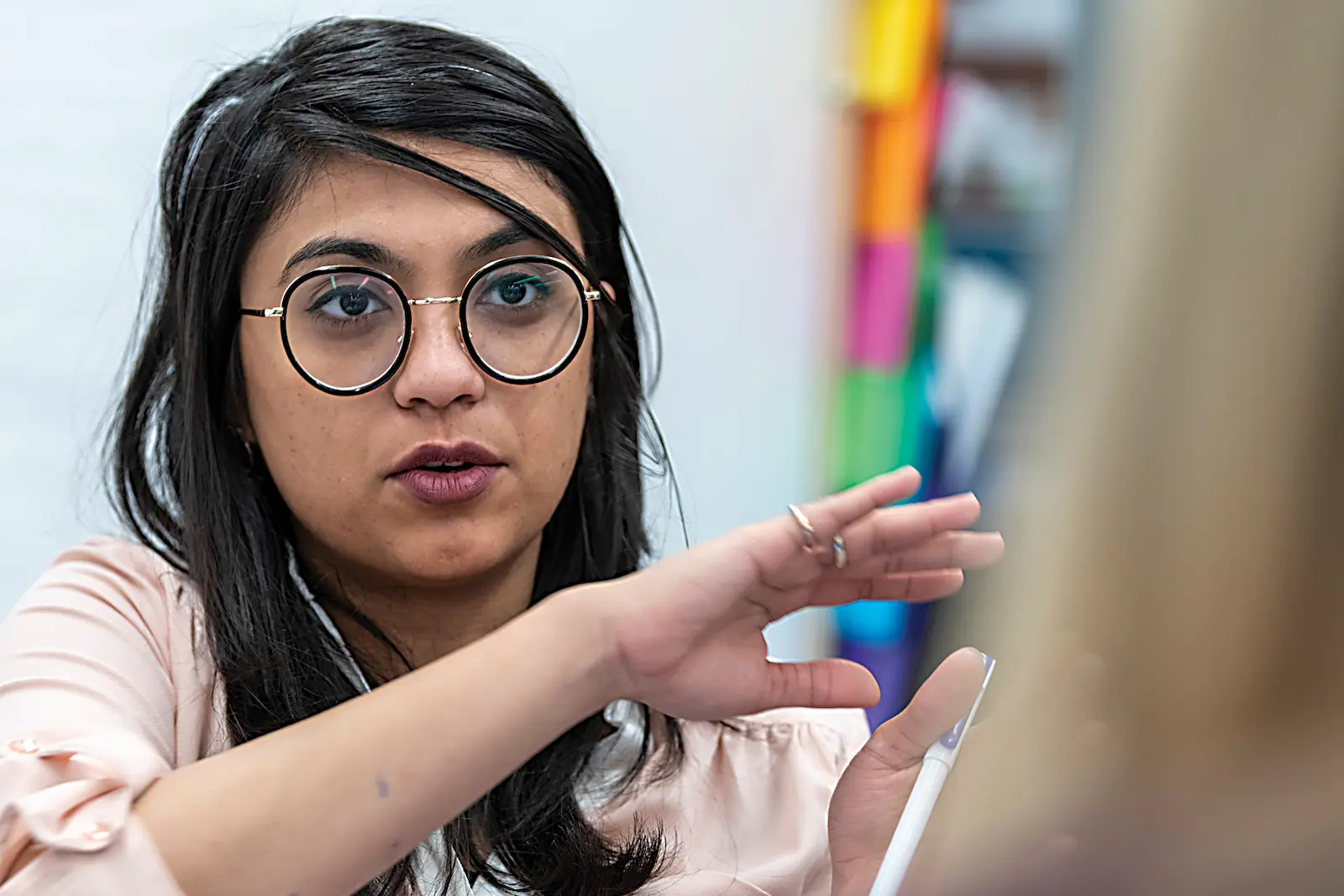 A student with dark hair, wearing round glasses and a pale pink shirt, gestures with her hands while speaking in class.