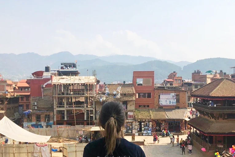 A person with a ponytail, seen from behind, looks over a townscape with traditional and modern buildings, some appearing to be under construction.  Rolling hills are visible in the background.