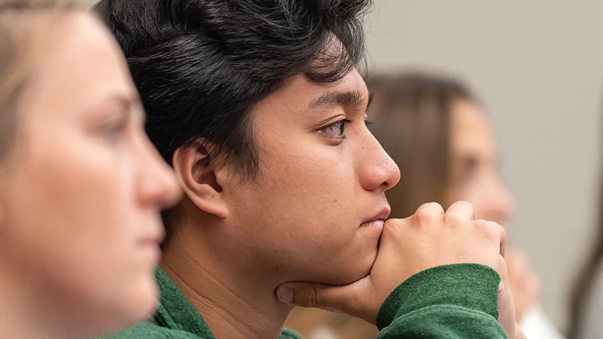 A student with dark hair, wearing a green sweatshirt, rests their chin on their hands and listens attentively in class.