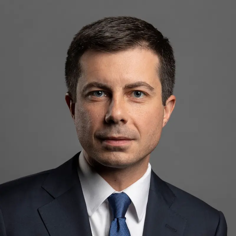 Pete Buttigieg, with short dark hair and blue eyes, wears a navy suit and blue tie, looking directly at the viewer.