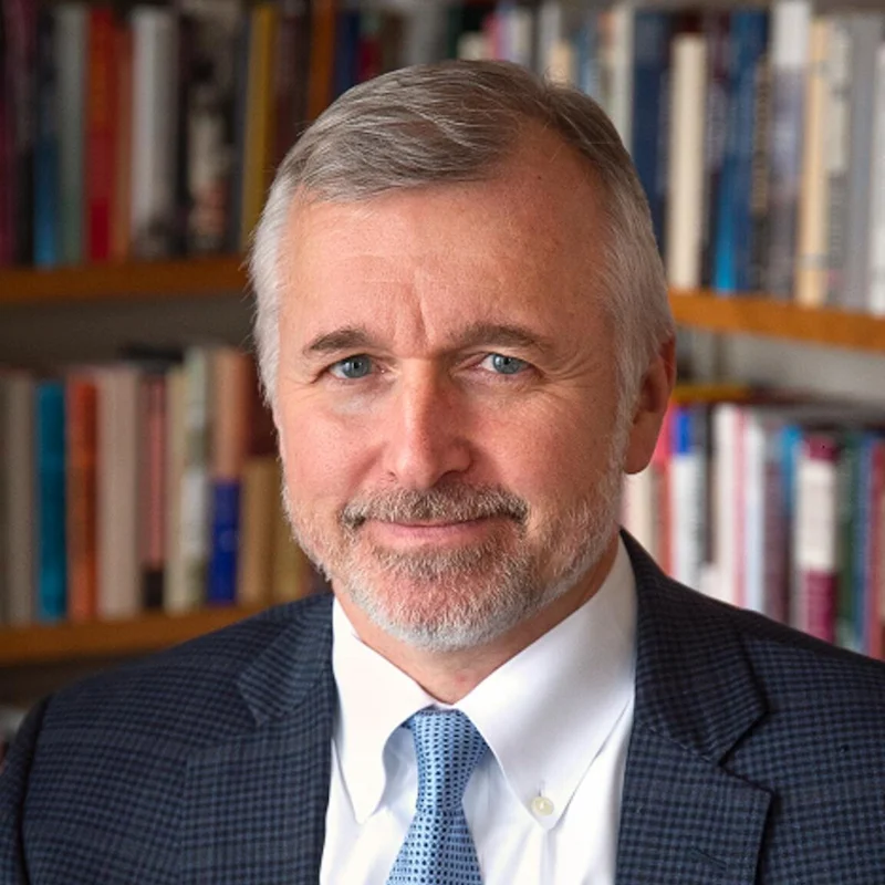 Thomas Christensen, a man with short grey hair, a grey beard, and blue eyes, wearing a white shirt, blue patterned tie, and dark checkered blazer, smiles gently in front of a blurry bookshelf.