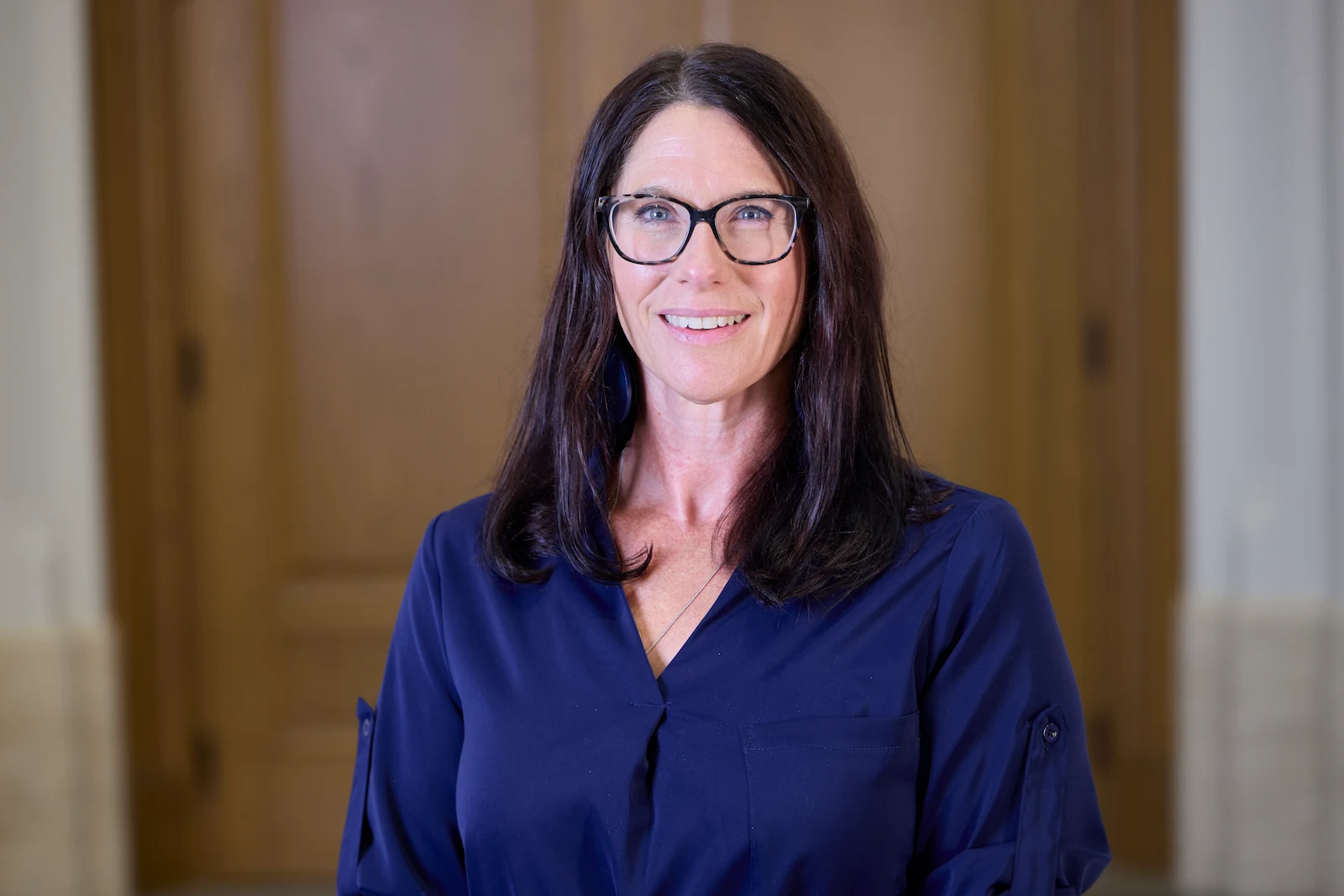 Tracy Kijewski-Correa, a smiling woman with dark hair and black glasses wears a navy blue shirt in front of blurry wooden doors.