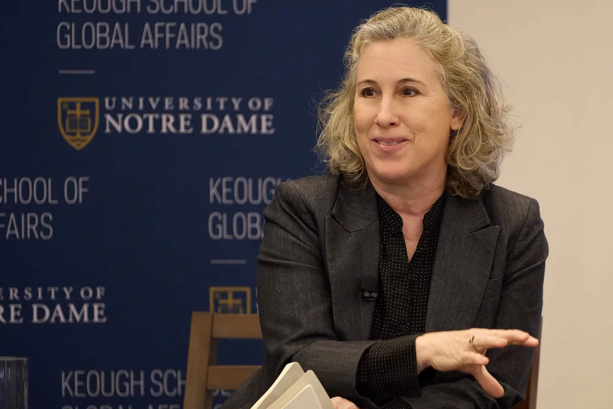 Smiling woman with curly blonde hair in a gray blazer speaks, gesturing, in front of the Keough School of Global Affairs banner.