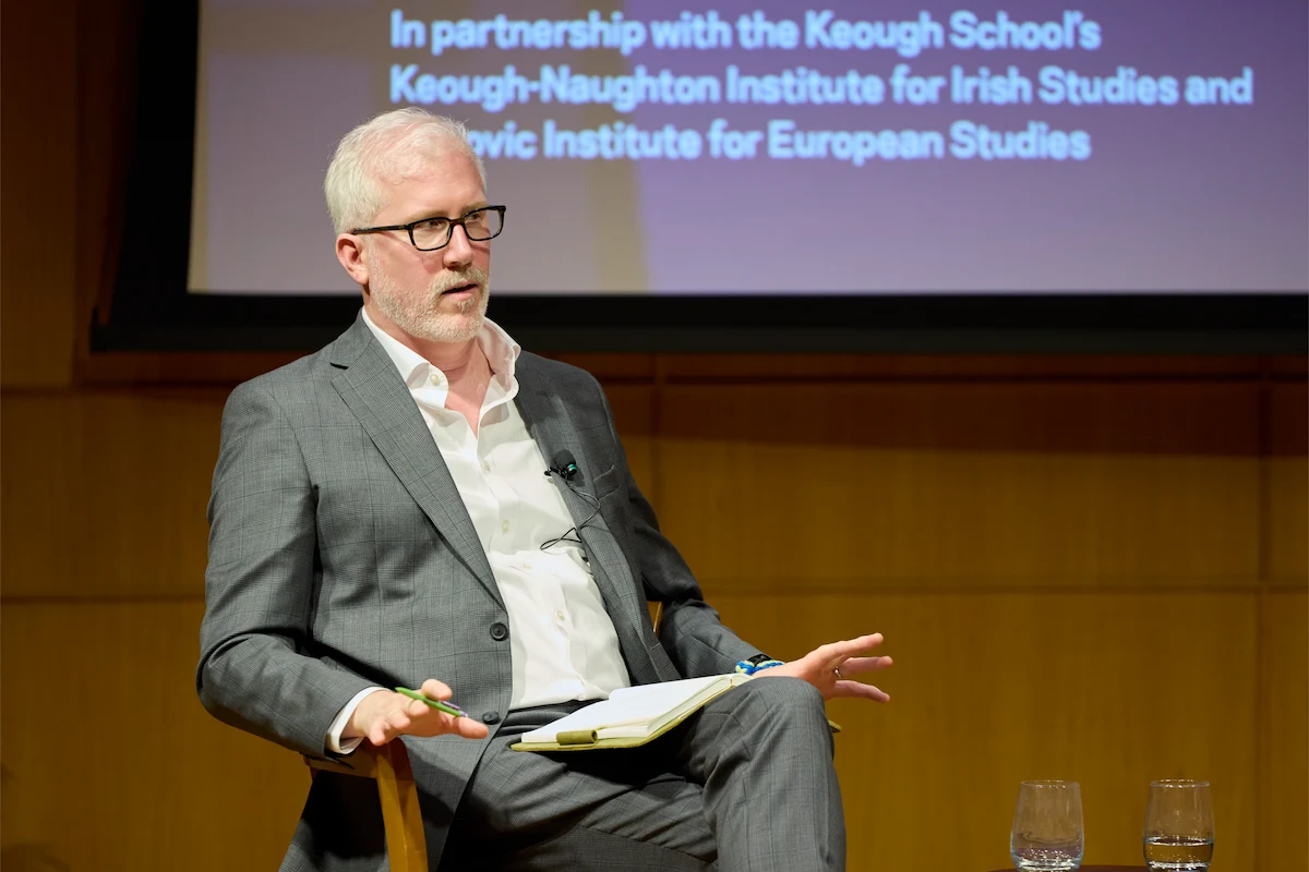 Rory Finnan, a white-haired man in grey suit, glasses speaks while holding a pen and notebook. Screen shows Keough-Naughton Institute.