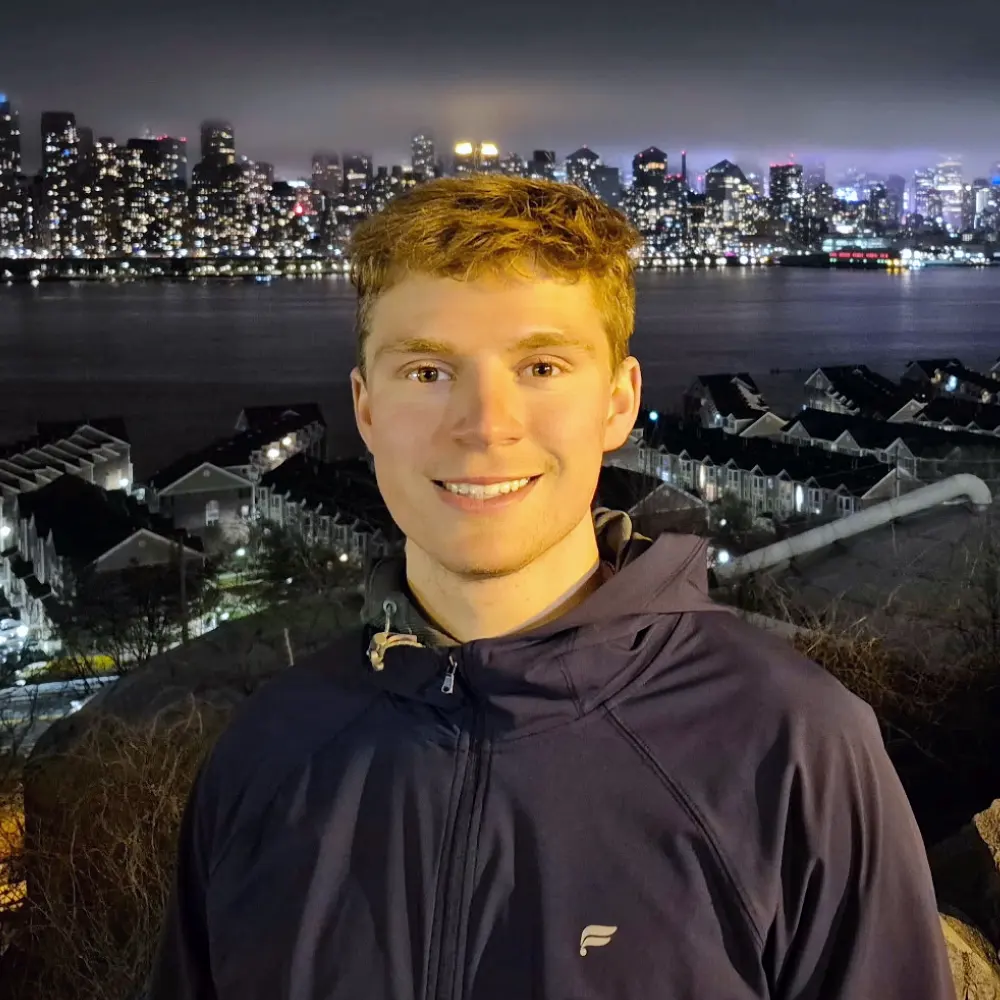 Patrick Marshall, a smiling young man with reddish-blonde hair in a navy jacket stands before a shimmering night city skyline over water.