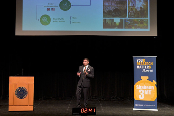 A speaker in a dark suit with a striped tie presents at the Notre Dame Shaheen 3MT competition. A timer reads 02:41.