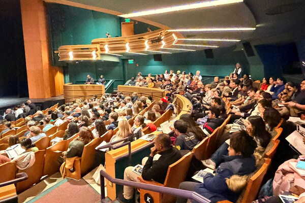 A large audience fills an auditorium with curved wooden seating and green walls. Many people face forward, some hold papers.
