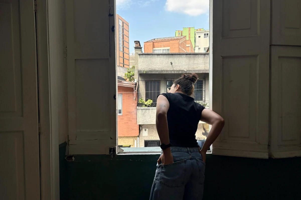 A person with a bun, in a black shirt and jeans, looks out an open window at multi-colored city buildings against a blue sky.