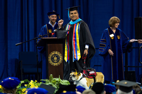 Smiling graduate in black gown, colorful stole, pumps fist on Notre Dame stage with bulldog in red service vest and cap.