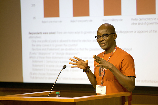 A man in an orange shirt and glasses, speaks and gestures at a podium before a screen with survey data.