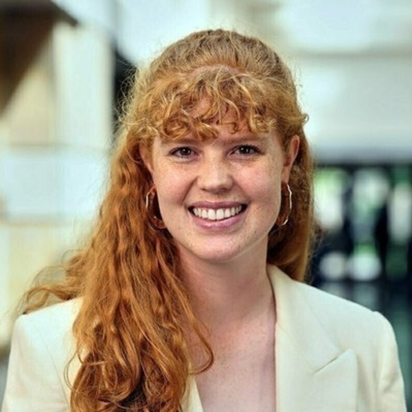 A smiling woman with long curly red hair, freckles, wearing a cream blazer and geometric gold hoop earrings.