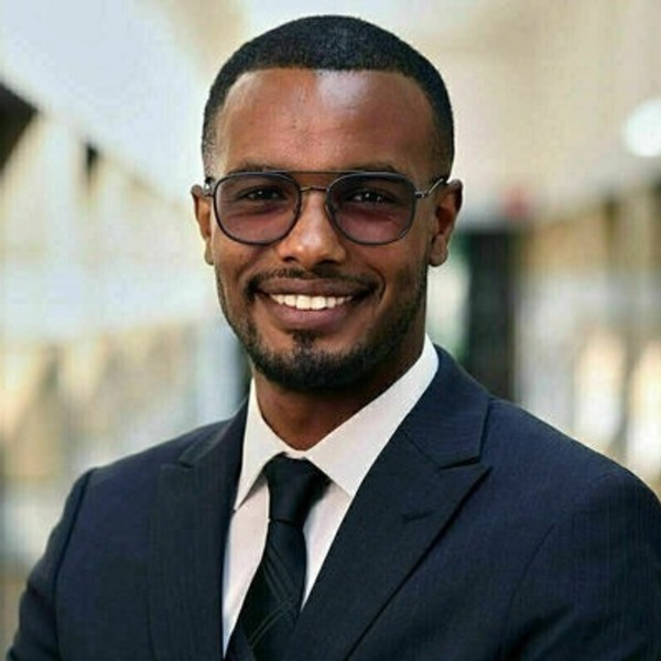 Smiling man with dark skin, short hair, beard, and dark glasses, wearing a dark suit, white shirt, and tie.