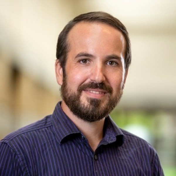 A man with a dark beard and dark hair wearing a purple striped shirt smiles directly at the camera.