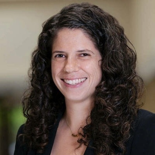 A woman with long, dark, curly hair smiles broadly at the viewer, wearing a dark blazer.