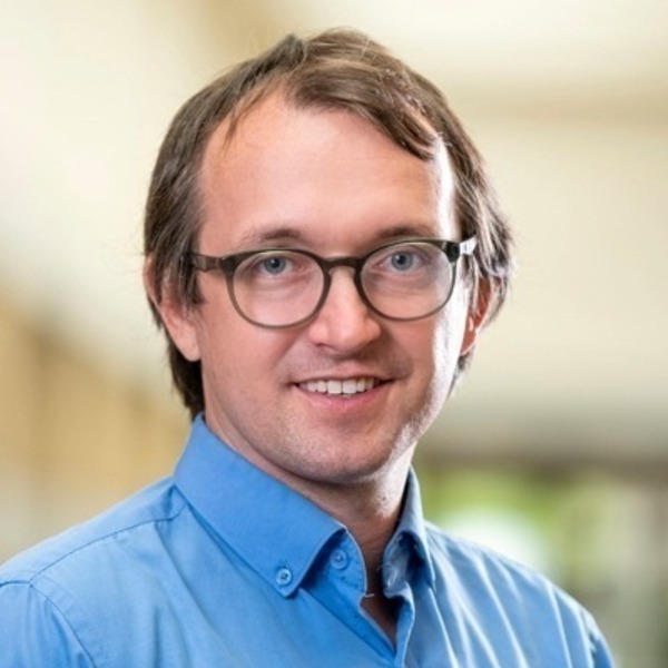 A man with light brown hair and dark-rimmed glasses, wearing a blue button-down shirt, smiles.