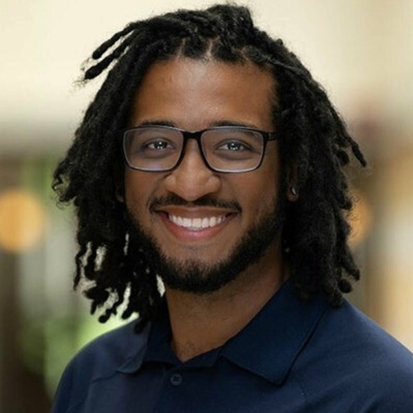 Smiling man with dark skin, black dreadlocks, glasses, and a beard wearing a dark blue polo shirt.