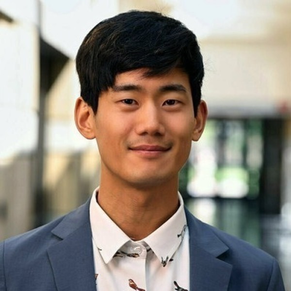 Young man with dark hair and a gentle smile, wearing a blue blazer over a white shirt with bird patterns.
