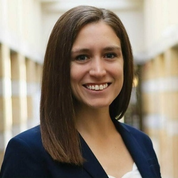 A smiling woman with shoulder-length brown hair, wearing a dark blue blazer and a white top, stands in a blurred hallway.