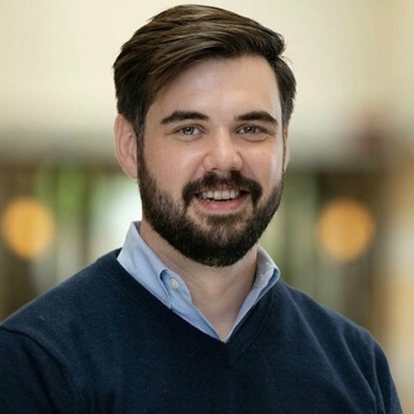 A smiling man with dark hair and beard wears a light blue shirt and navy V-neck sweater.