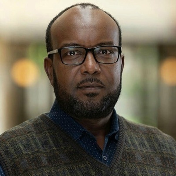 Dark-skinned man with beard and glasses wears a blue patterned shirt and a dark knitted vest, looking forward.