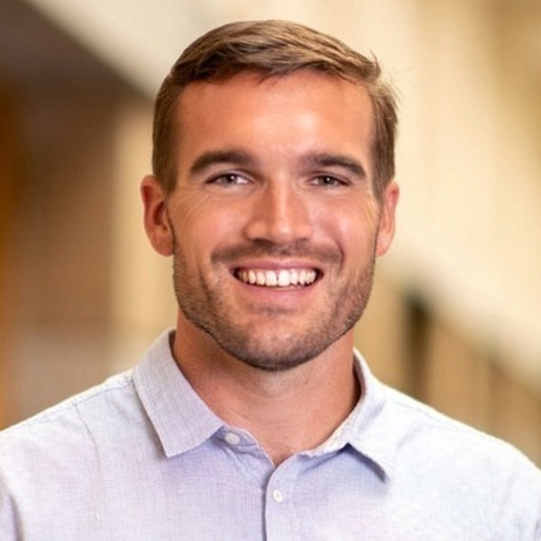 A young man with short brown hair and light stubble smiles broadly, wearing a light blue button-up shirt.