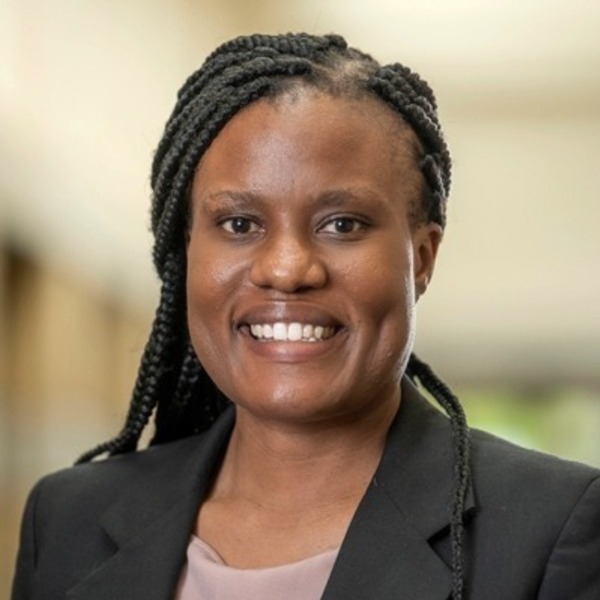 A Black woman with dark braided hair smiles, wearing a black blazer and light pink top.