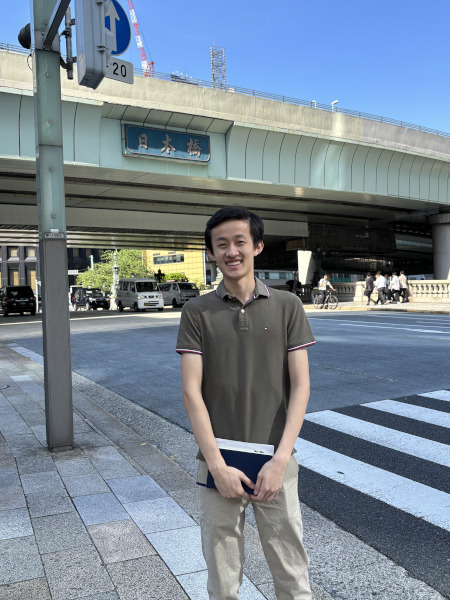 A young man smiles, wearing an olive polo and khaki pants, standing by a crosswalk under the Nihonbashi sign.