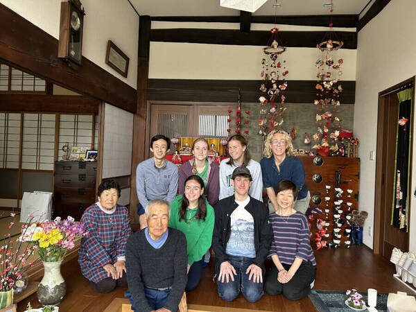 Nine people, including two elderly Japanese hosts, smile in a traditional Japanese home with wooden floors and cultural decorations.