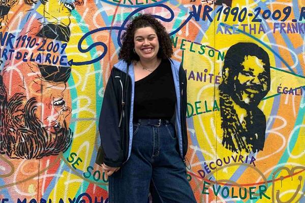 A smiling woman in a blue jacket and jeans stands against a vibrant graffiti on the Berlin Wall that features Nelson Mandela and Aretha Franklin.
