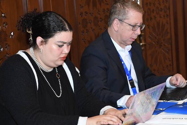 A focused woman wearing pearl necklaces and a man in a suit work at a table with laptops.