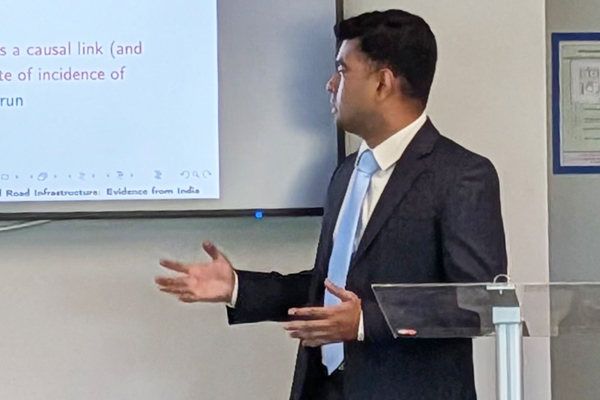 Man in a dark suit and light blue tie gestures at a screen displaying a research presentation.