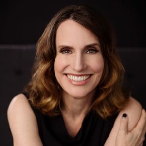 Smiling woman with brown wavy hair and brown eyes wearing a black sleeveless top against a dark background.