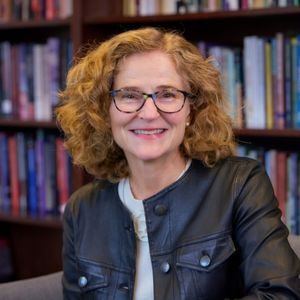 Smiling woman with glasses, curly red-brown hair, and a black jacket sits in front of bookshelves.