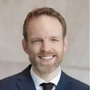 Smiling man with light brown hair and beard wearing a dark suit and tie.
