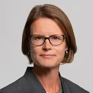 Woman with short brown hair and glasses smiles faintly, wearing a grey blazer and a silver necklace.