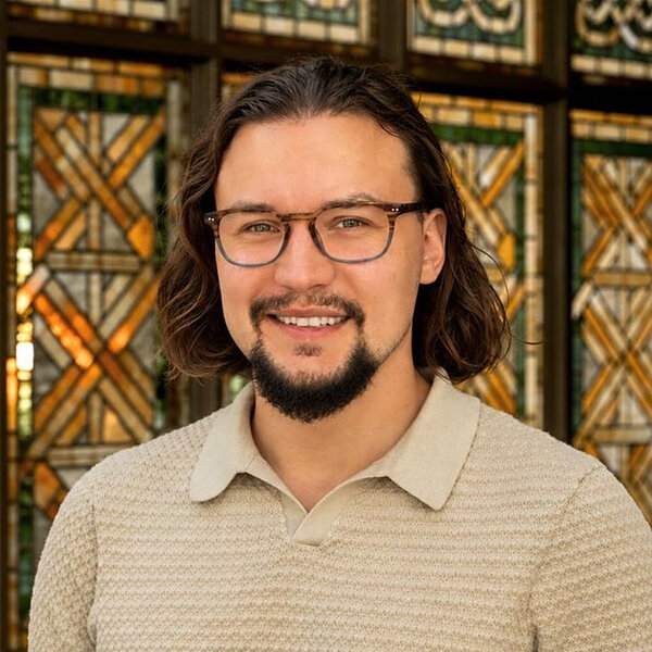 Man with brown hair and glasses smiles in front of stained glass windows.