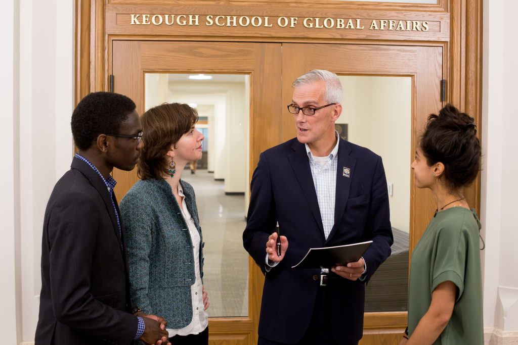 Denis McDonough speaking to three students in front of the entrance to the Keough School Dean's Office